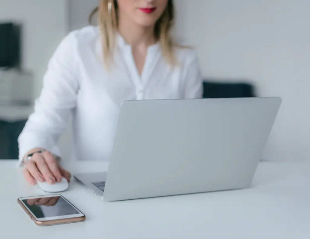 Frau arbeitet am Laptop, daneben ein Smartphone auf einem weißen Schreibtisch, für den Service der Digital Agency Jinni S.r.l.