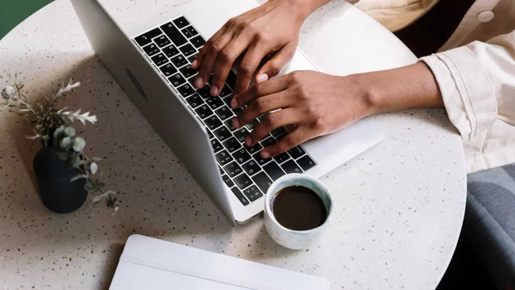 Hombre tecleando en un portátil con un café al lado, sobre una mesa redonda, escribiendo contenidos para la web