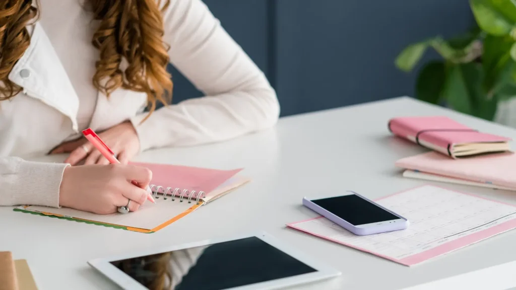 Mulher a escrever num caderno, com planner e dispositivos digitais em cima da mesa, para o guia dos principais tipos de conteúdos para as redes sociais.