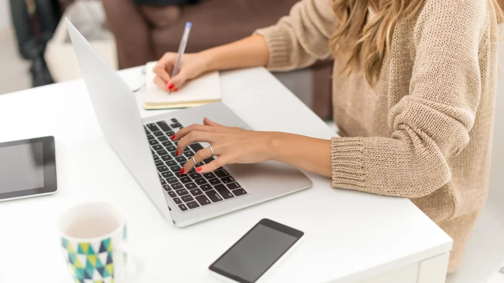 Frau, die an einem Laptop schreibt und sich auf einem Notizblock Notizen macht, für einen Artikel über das lokale Geschäft im Web.