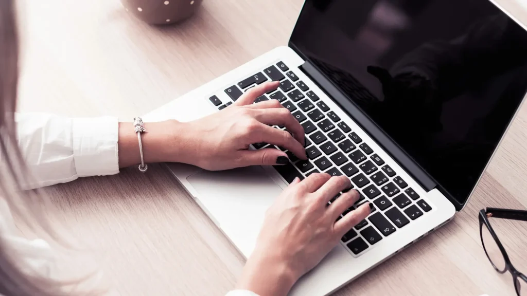 Frau, die an einem Laptop auf einem hellen Schreibtisch schreibt, mit einem Armband am Handgelenk und einer Brille daneben, für einen Artikel über das lokale Geschäft im Web.
