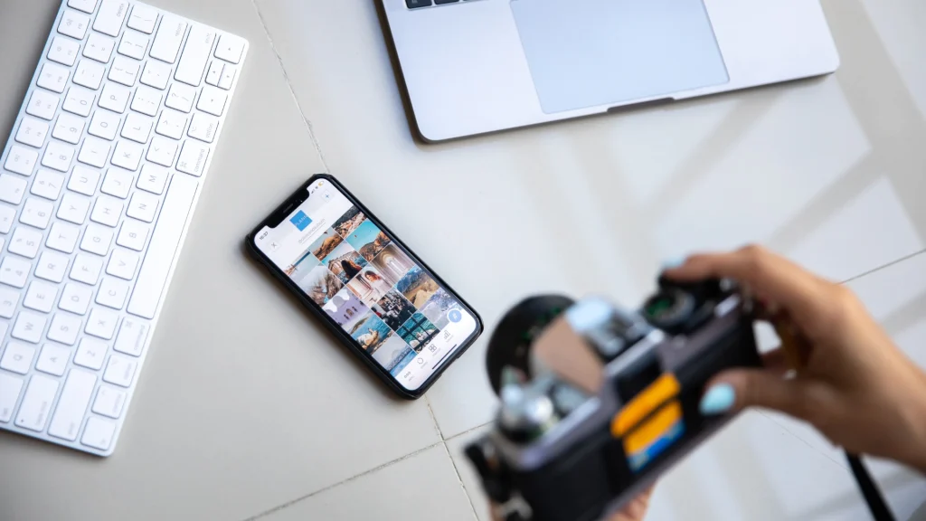 Smartphone apoyado en un escritorio que muestra una galería de imágenes en cuadrícula, junto a un teclado y una cámara, para el artículo sobre formatos de redes sociales