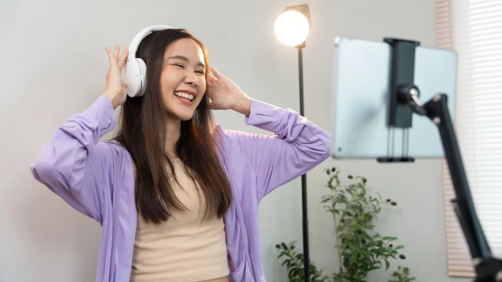 Mujer sonriente con auriculares blancos que graba un reel con un smartphone montado en un soporte, para el artículo sobre formatos de redes sociales