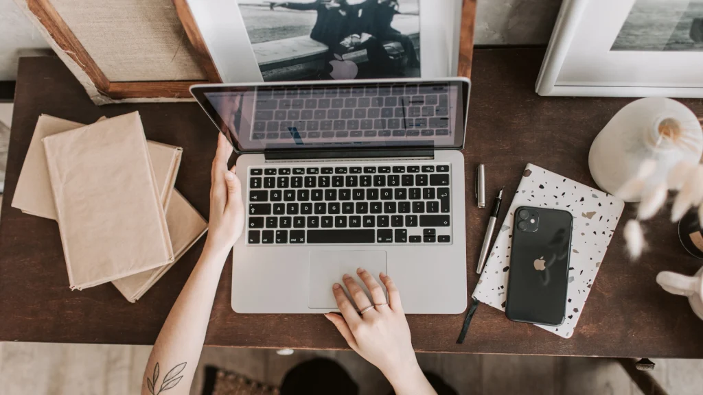 Person öffnet einen Laptop auf einem Holzschreibtisch, daneben Smartphone, Notizbuch, Stift und Bücher.
