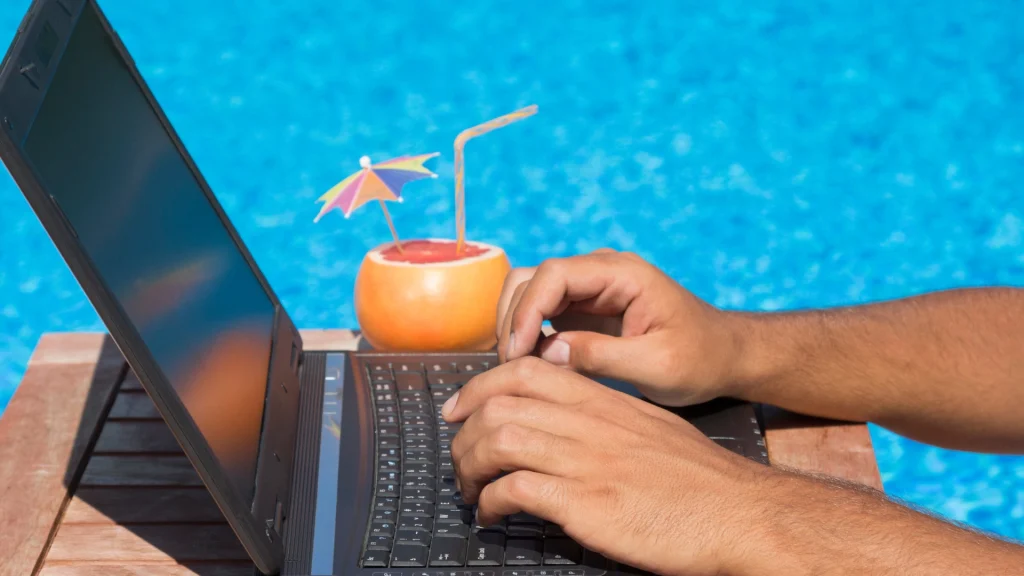 Hands of a man typing at laptop by the pool with tropical cocktail next to