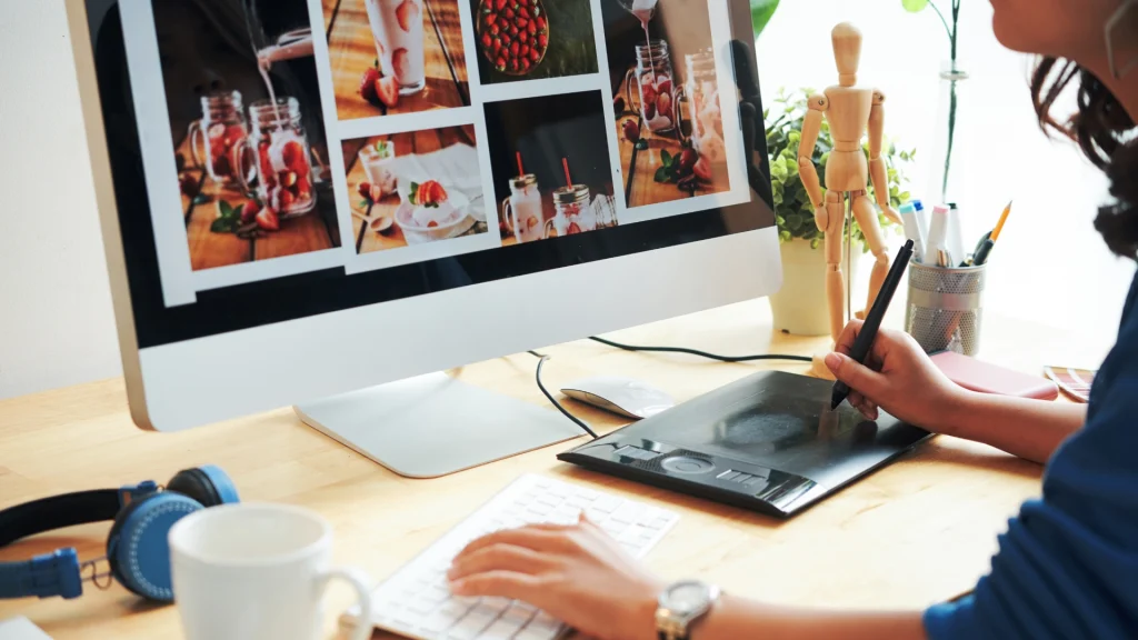 Person working on a digital graphic design on the computer with graphic tablet, photographs and design tools on the desk.