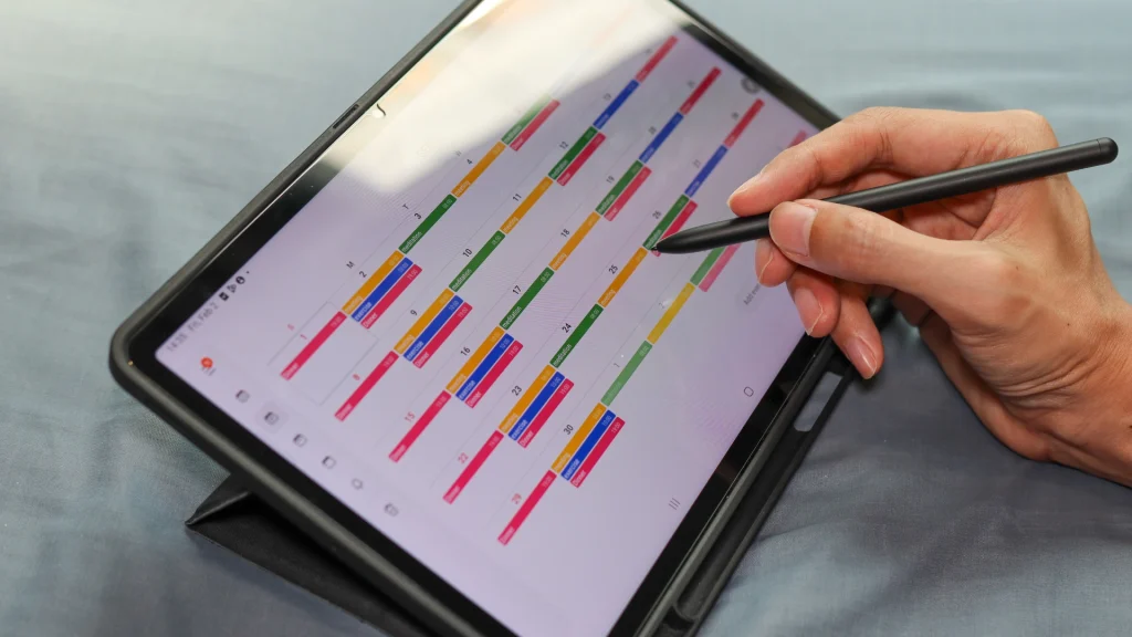 Hand using a digital pen on a tablet with colorful weekly calendar in the foreground.