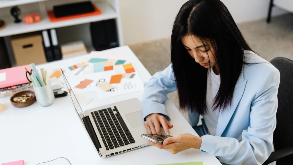 Woman uses smartphone while working on laptop in office