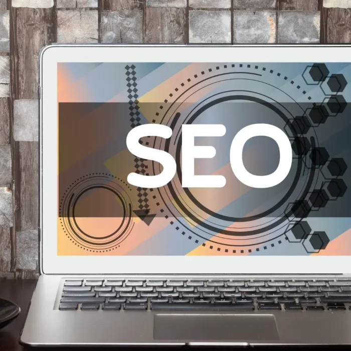 Schermo di un laptop con la scritta “SEO” su sfondo grafico colorato, accanto a una pianta e una tazza per l'articolo su come migliorare il posizionamento del sito web