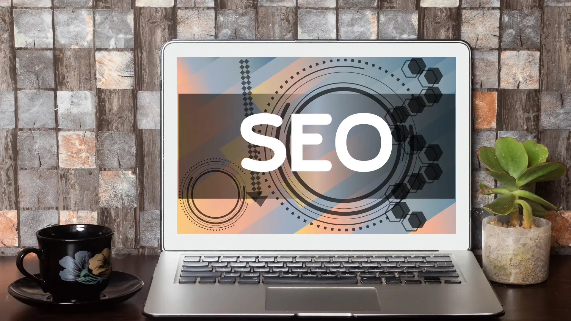 Schermo di un laptop con la scritta “SEO” su sfondo grafico colorato, accanto a una pianta e una tazza per l'articolo su come migliorare il posizionamento del sito web