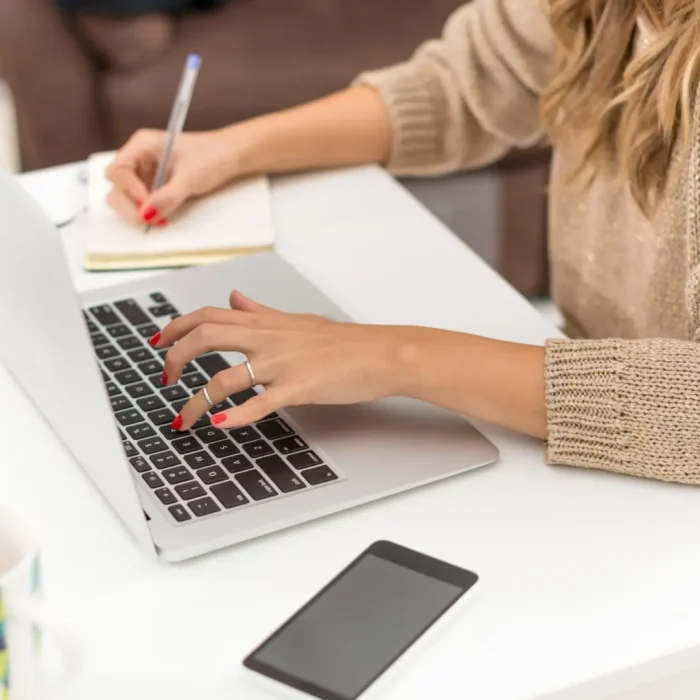 Woman writing on a laptop and taking notes in a notebook for article dedicated to local activity on the web.