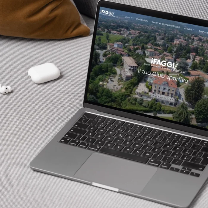Laptop aperto sul sito web iFaggi Biella, poggiato su divano con AirPods accanto.