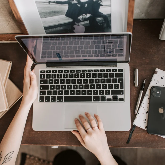 A person opening a laptop on a wooden desk, with a smartphone, notebook, pen, and books.