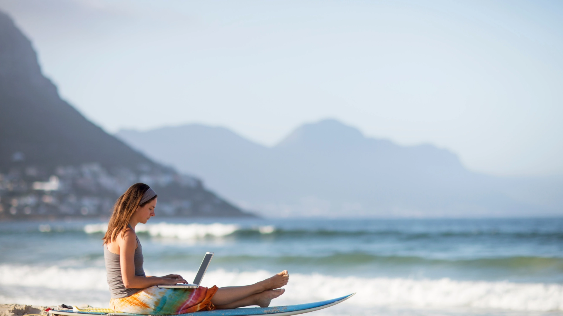 Donna lavora con il laptop su una tavola da surf davanti al mare, rappresentando il potere delle automazioni anche in ferie.
