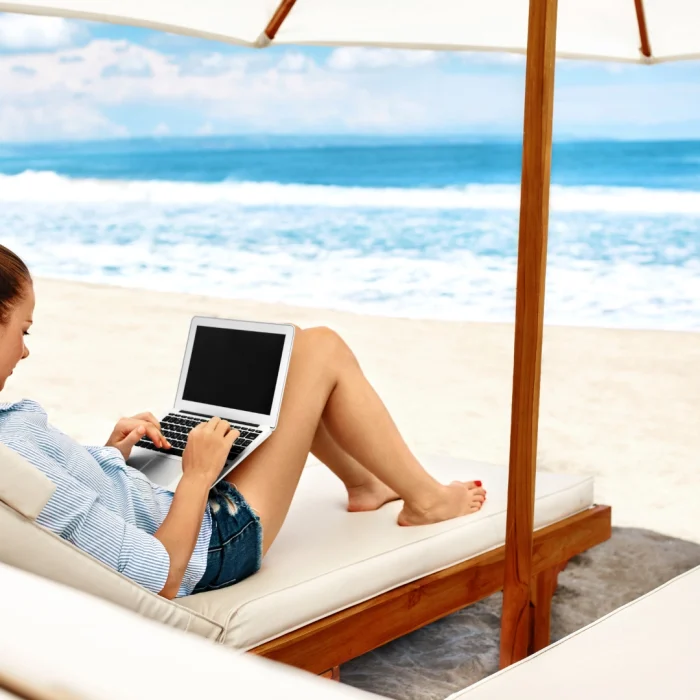 Donna che segue un corso online al computer, sdraiata su lettino in spiaggia sotto un ombrellone, con vista mare