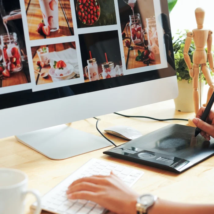 Person working on a digital graphic design on the computer with graphic tablet, photographs and design tools on the desk.