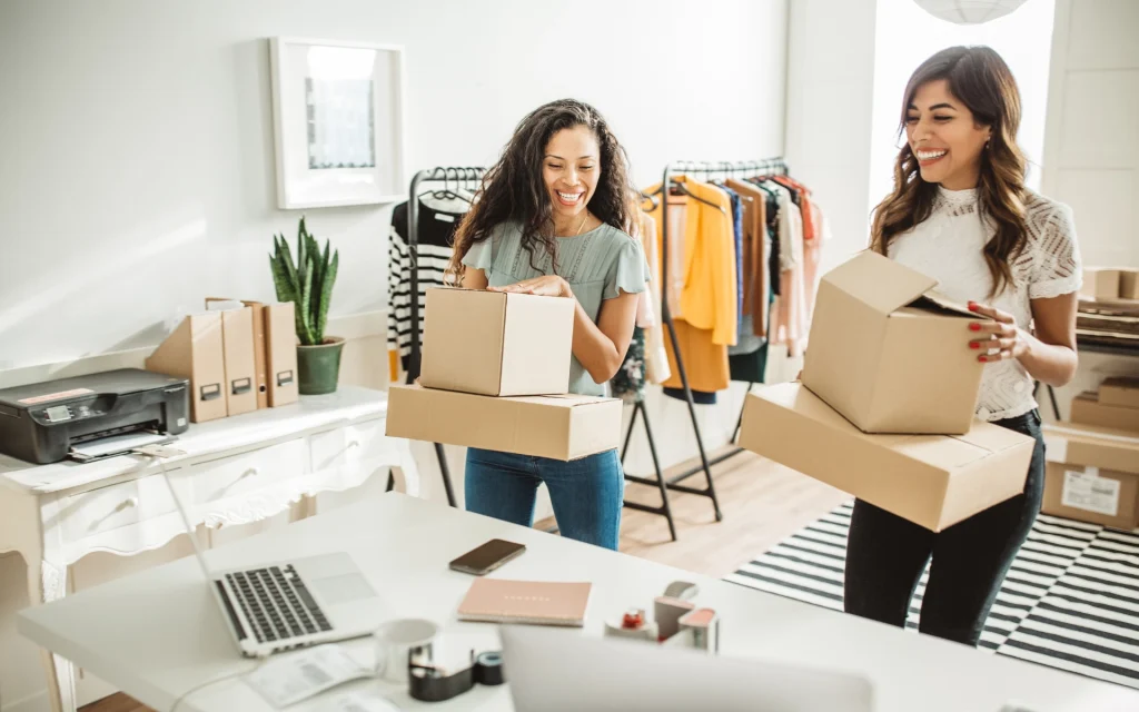 Due donne in un piccolo magazzino di moda: una prepara pacchi su un tavolo con laptop, l’altra sistema capi d’abbigliamento su appendiabiti.
