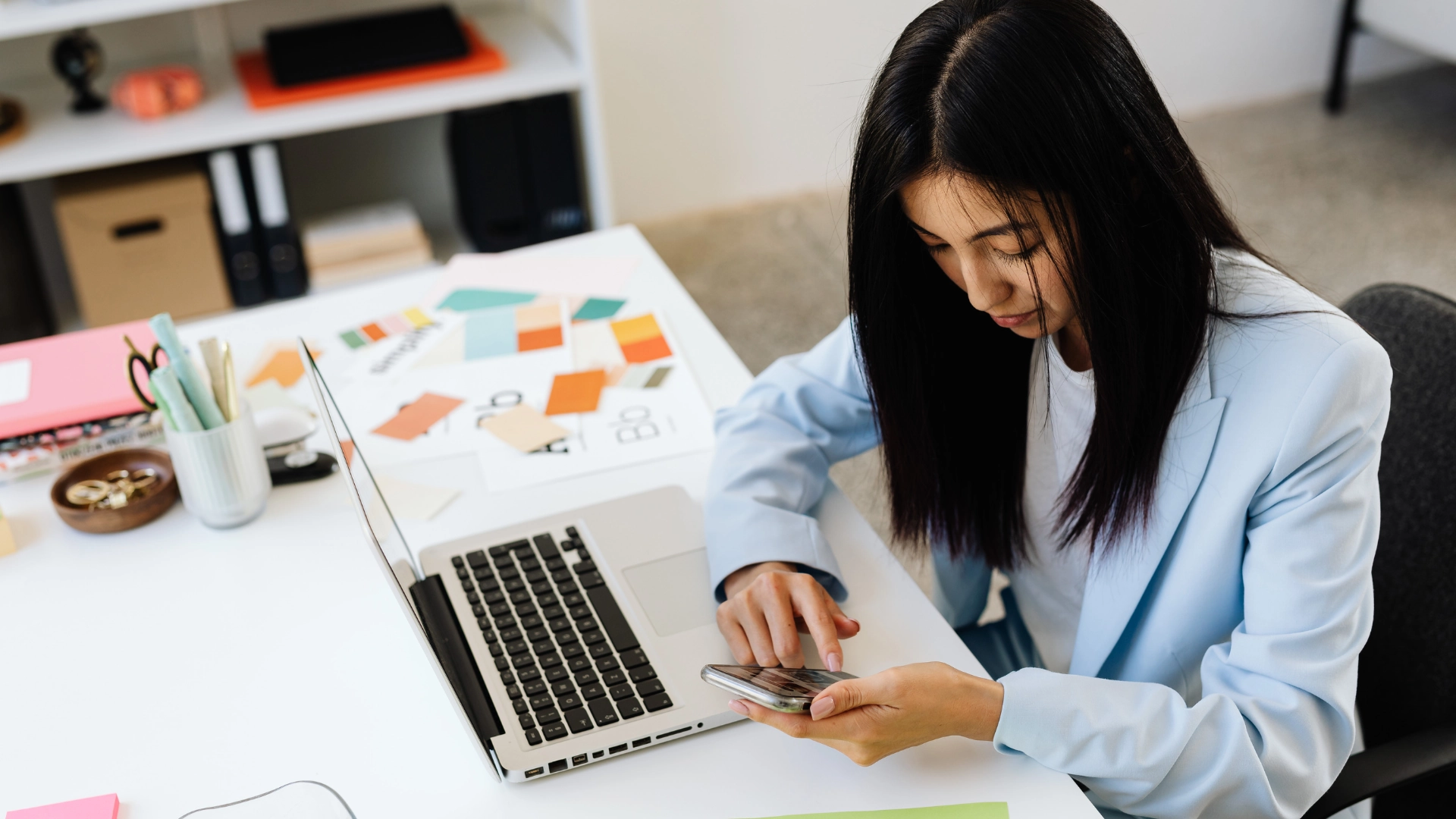 Woman uses smartphone while working on laptop in office